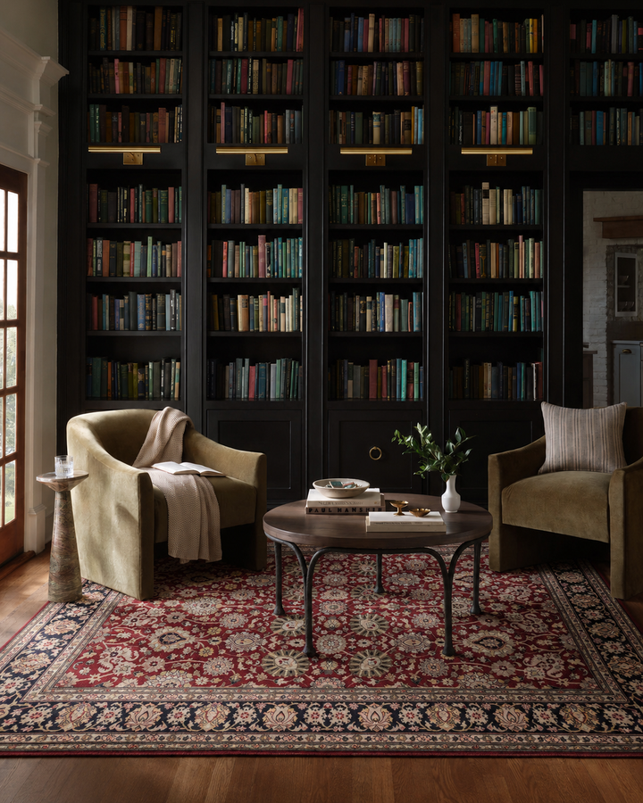 A cozy library reading nook with tall black bookshelves filled with books, two olive green armchairs, a round wooden coffee table, and a large red and navy patterned area rug on a hardwood floor.